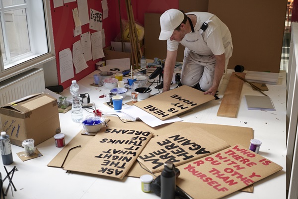 A man kneeling on the ground painting a placard with the words 'here is my home'