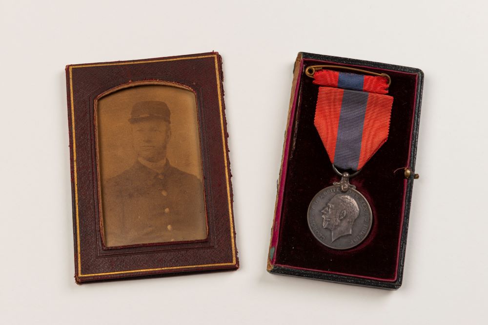 On the left, a framed photo of Albert Hussey in uniform. On the right, his Imperial Service Medal. The medal has a red ribbon with a navy stripe down the middle, and the medal is silver. 