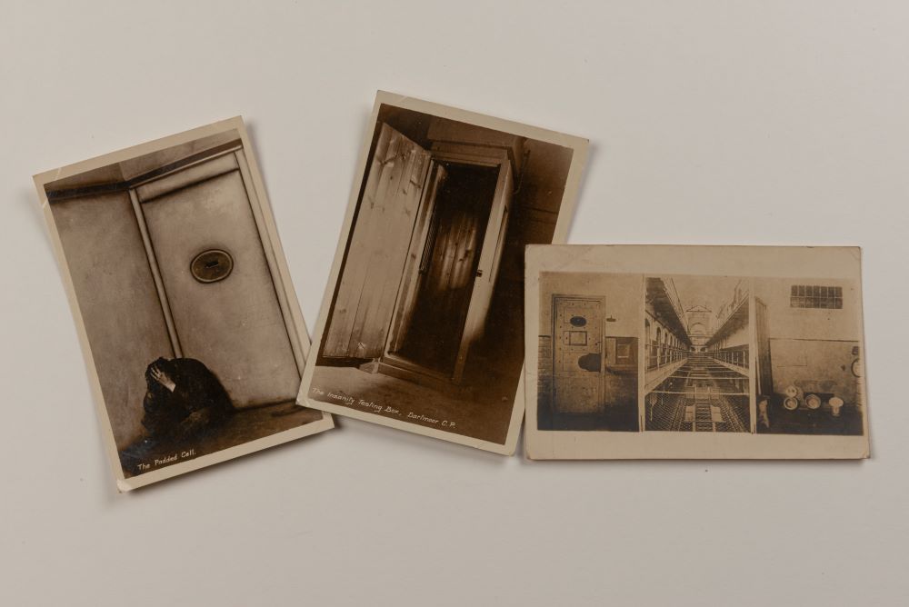Three postcards of Dartmoor prison. The first shows a prisoner sitting in the corner of a padded cell, head in their hands. The middle is of a very narrow cupboard, captioned 'the insanity testing box'. The final is a collage of images of the interior of the prison. 