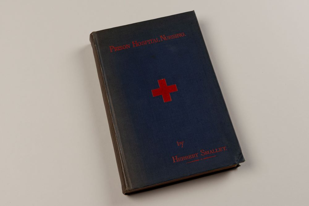 The front cover of Prison Hospital Nursing, the book is navy with a red cross and red title and author. 