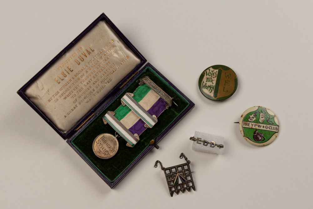 Elsie Duval's hunger strike medal, with the suffragette colours in the ribbon. The medal is within its case, with her name inscribed. The case is surrounded by other badges Elsie had owned.