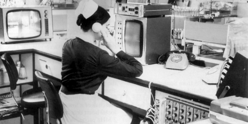 Greyscale image of a nurse seated at a desk surrounded by various boxy monitors and technological items with knobs and buttons. She has her back to the camera and is holding the handset of a rotary phone to her ear.