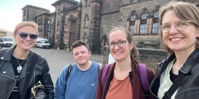 Landscape image of the RCN Museum team, four smiling white people, 3 women and 1 man, standing in front of the grand stone front of HMP Wandsworth.
