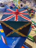 A 'memory box', a hand-decorated box featuring a red, white and blue design resembling the flag of Great Britain on t he top and a green, yellow and black design resembling the Jamaican flag on the front. The designs are embellished with sequins and glitter. Various crafting materials are visible in the background including pens, twine, foam stickers and feathers.