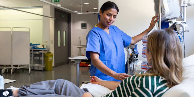 A nurse stands talking to a woman patient who is in a bed. She is taking a reading and smiling at the patient. The nurse has dark hair and wears a blue uniform and the woman in bed has blonde hair and is wearing a green and white striped top, grey trousers and trainers