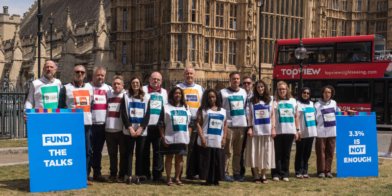 Members representing different health unions are shown standing on a patch of grass in Westminster. They are wearing white shirts with various logos and colours of the unions on them. Two placards read "fund the talks" and "3.3.% is not enough". A red London bus is seen behind them.