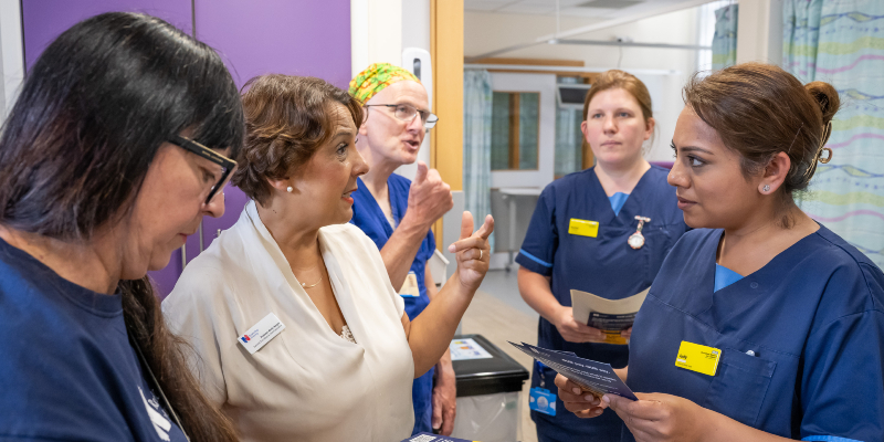 A group of nursing staff in blue uniforms stand gathered in a hospital. Among them, the RCN Chief Executive and General Secretary Professor Nicola Ranger talks to them about pay. They are holding leaflets and look deep in conversation. Nicola stands in the middle and wears a cream coloured top