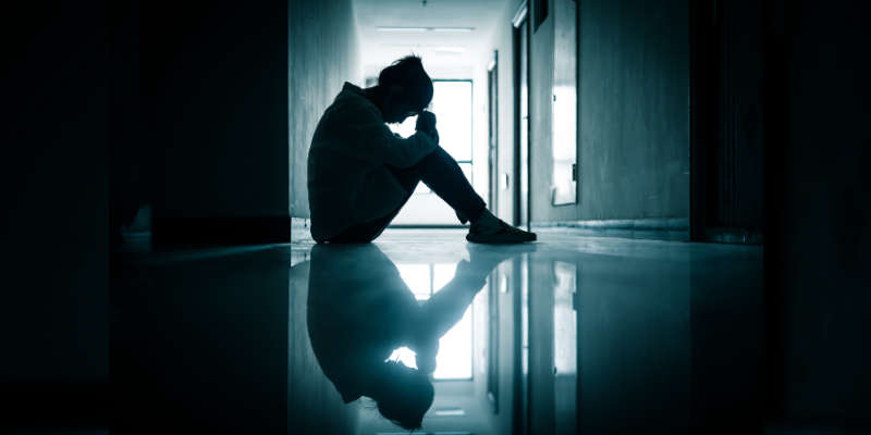 A silhouetted figure sits in a dark hallway with their reflection visible in a shiny floor below them. They are facing to the right and the background has a black and blue colour. Their posture suggests sadness or stress.