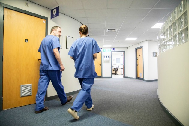 Two members of nursing staff walk down a corridor. One is male and one female. They both wear blue uniforms and are walking away from the camera, from the left to the right. Wooden doors can be seen off the corridor in the foreground and background, as the corridor bends around to the right