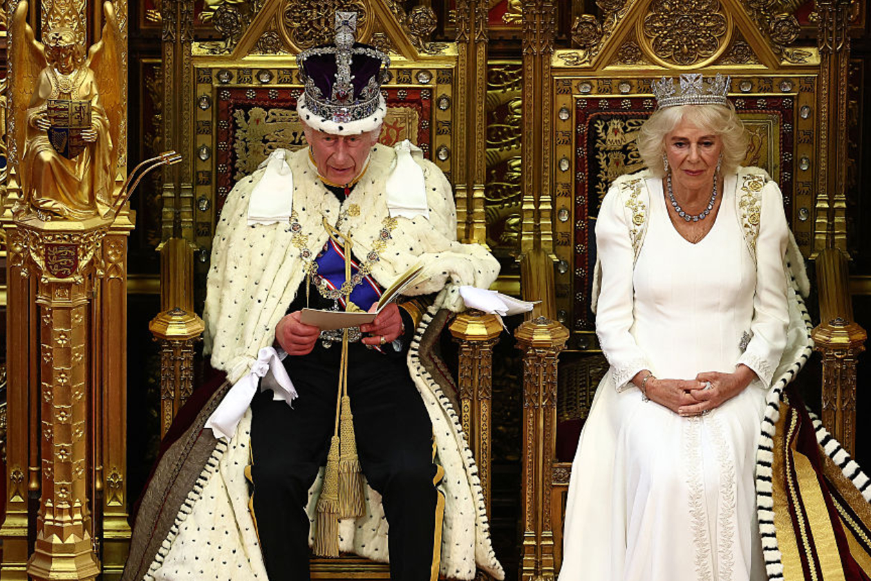 King Charles and Queen Camilla sit on golden thrones wearing crowns and official royal garb, at the King's Speech on 13 May 2026. The King wears black trousers and shoes with cream robes and a purple sash. He is reading from a booklet. The Queen wears all white and sits with her hands crossed in her lap.