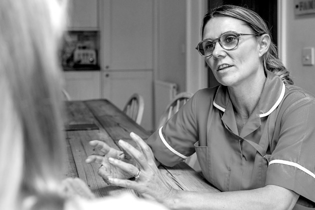 Black and white photo shows district nurse sitting at table speaking to patient
