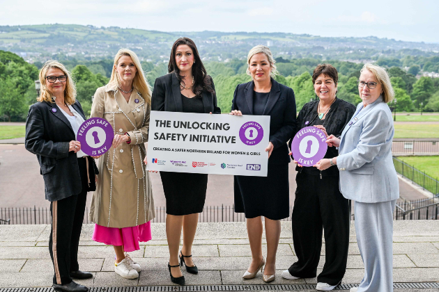 A group of women stand together outside at Stormont holding a banner and signs reading "Unlocking Safety Initiative". The RCN's Rita Devlin stands second from the right, along with First Minister Michelle O’Neill, Deputy First Minister Emma Little-Pengelly and representatives from Women's Aid. Hills covered in greenery can be seen behind them, below a cloudy sky.