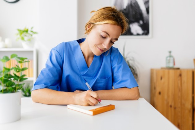 Nurse in blue uniform sat at a table  writing in a notepad.