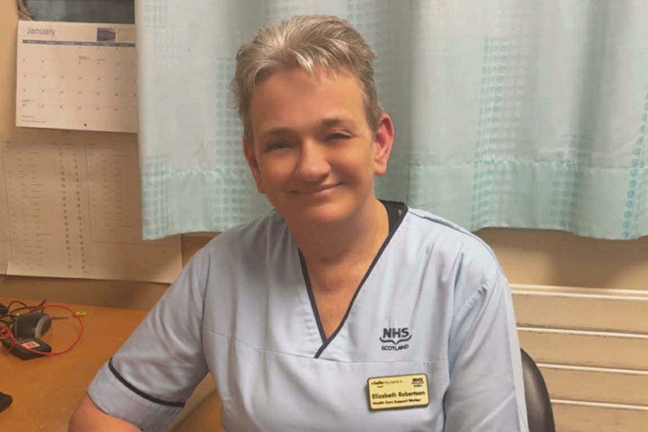A health care support worker is shown, sitting and smiling while looking directly into camera. She wears a light blue uniform with a yellow name badge. Her hair is cropped short. She is sitting inside, with her arm resting on a desk and curtains and a calendar visible behind her.