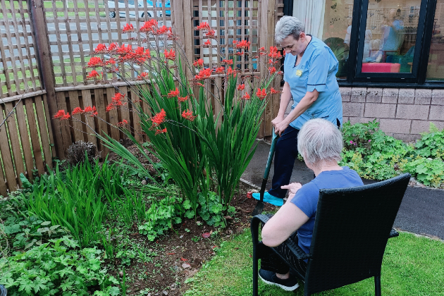 A garden is visible, with a health care support worker standing and digging, and a patient sitting in a black chair with their back turned towards the camera. Various plants are seen in a flower bed, including some red flowers, and a patch of lawn.