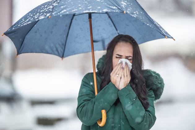 Person walking in rain with umbrella sneezing into tissue