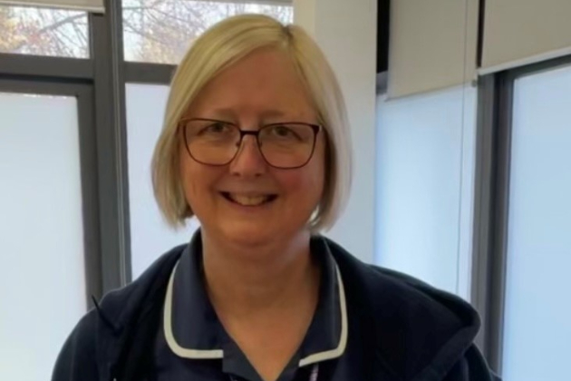 Alison, a district nurse, can be seen smiling and looking into the camera. She has blonde hair and glasses and is wearing her nursing uniform.