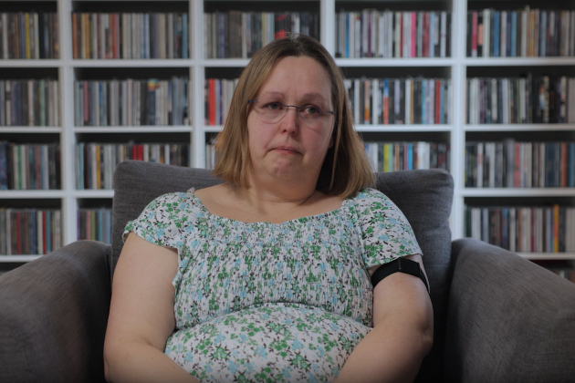 Lisa, a retired ICU nurse, is seen sitting in a large grey armchair, with shelves full of colourful CDs behind her. She has glasses, shoulder-length dark blonde hair, and is wearing a dress with blue and green flowers on it and looking off-camera. She is visibly emotional.