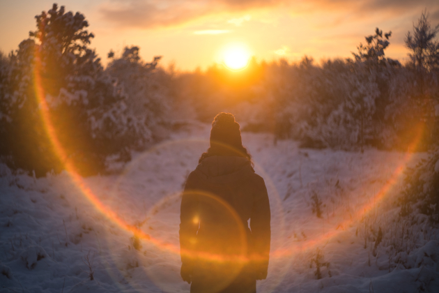 Woman wearing a winter coat, with her back to the camera, walking in snowy woodlands facing an orange rising sun