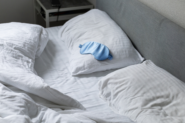 Unmade bed with white sheets and pillowcases, and a blue eye mask lying on one pillow
