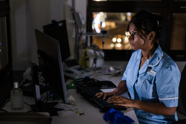 Female nurse of South Asian heritage wears blue nurse's uniform and sits at a computer in a hospital at night time