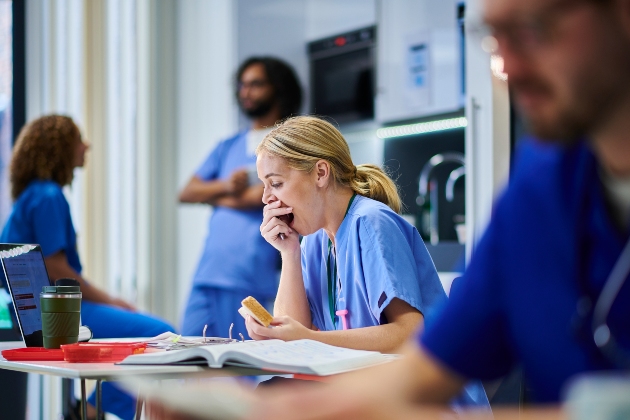 A white, blonde, young female nurse covers her hand with her mouth while she yawns. She is sitting at a table in a break room, with a sandwich, drink, laptop and paperwork.