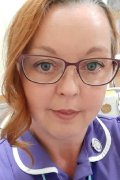 Michelle Ruecroft - close-up picture of a woman wearing dark-red-rimmed glasses with orangey-red hair tied back with a curl dangling down the right-hand side of her face and a purple uniform on with a badge on the left collar